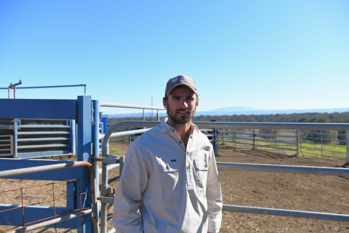 Man standing in front of a cattle yard smiling at the camera. He is wearing a white shirt and cap with his hands in his pockets.. 