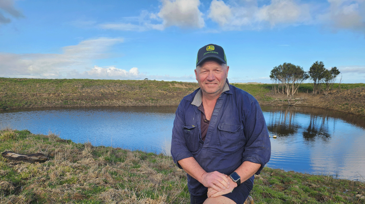 Smiling man wearing a blue cap, kneeling on grass in front of a blue dam.