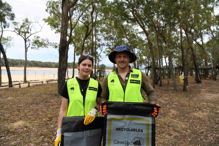 Two people standing at a lake with trees in the background holding rubbish bags smiling at the camera