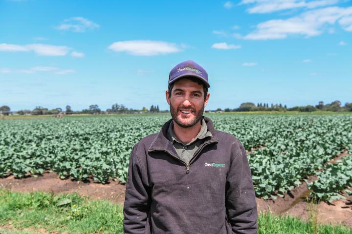 Man smiling at the camera wearing a cap and navy jumper standing in a vegtable farm.